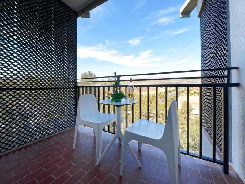 Balcón con dos sillas blancas y mesa pequeña con botella y vasos, vista al exterior.