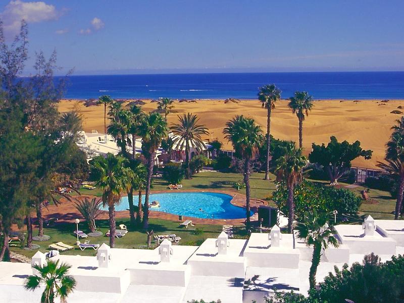 Hotel met zwembad, palmbomen en uitzicht op zee onder een heldere blauwe lucht.