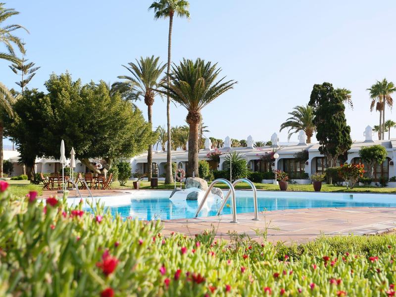 Zwembad in een hotel tuin met palmbomen, bloemen en parasols.