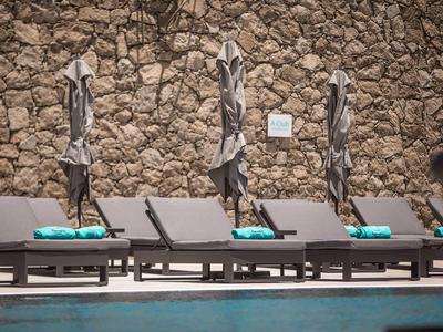 Three lounge chairs with closed umbrellas in front of a stone wall by a pool.