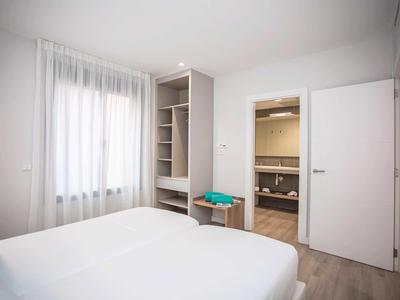 Dormitorio luminoso con cama blanca, ventana con cortinas y vista a baño con puerta abierta