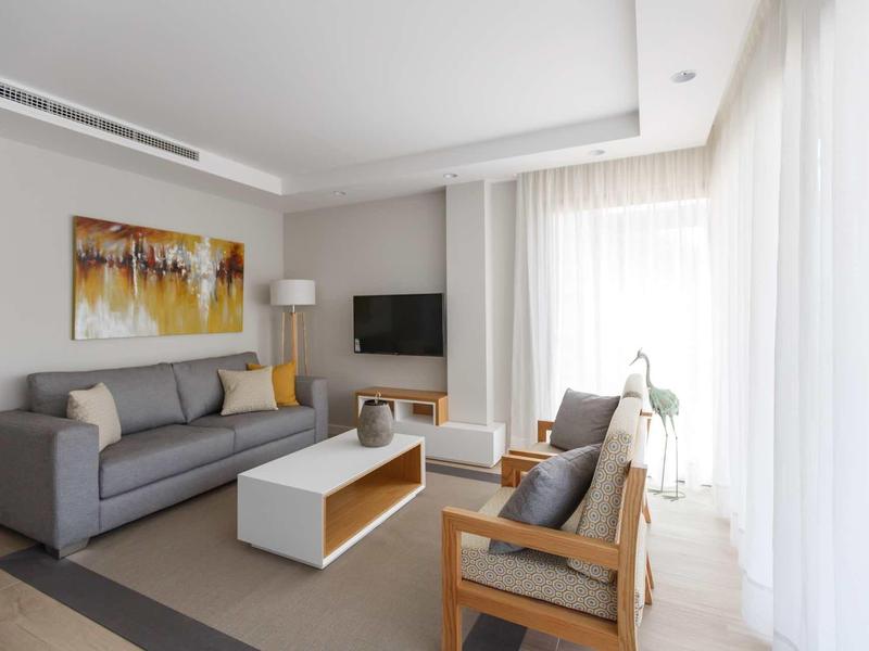 Sala de estar moderna con sofá gris, sillas de madera y ventana grande con cortinas blancas.