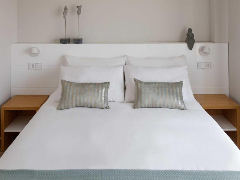 Dormitorio moderno con cama blanca, cojines grises y dos mesitas de noche de madera.