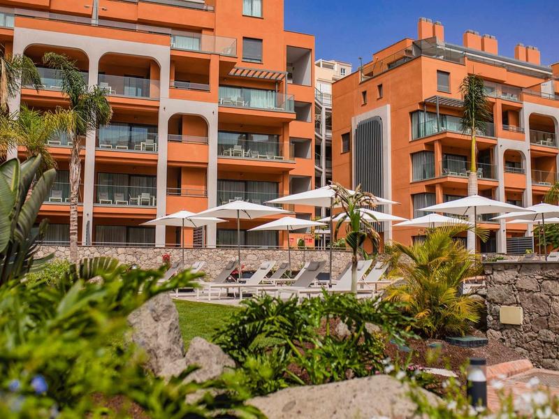 Complejo hotelero naranja con balcones, tumbonas y sombrillas frente a un muro de piedra con vegetación.