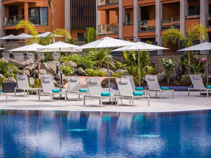 Piscina del hotel con tumbonas y sombrillas frente a un edificio de varios pisos con balcones.