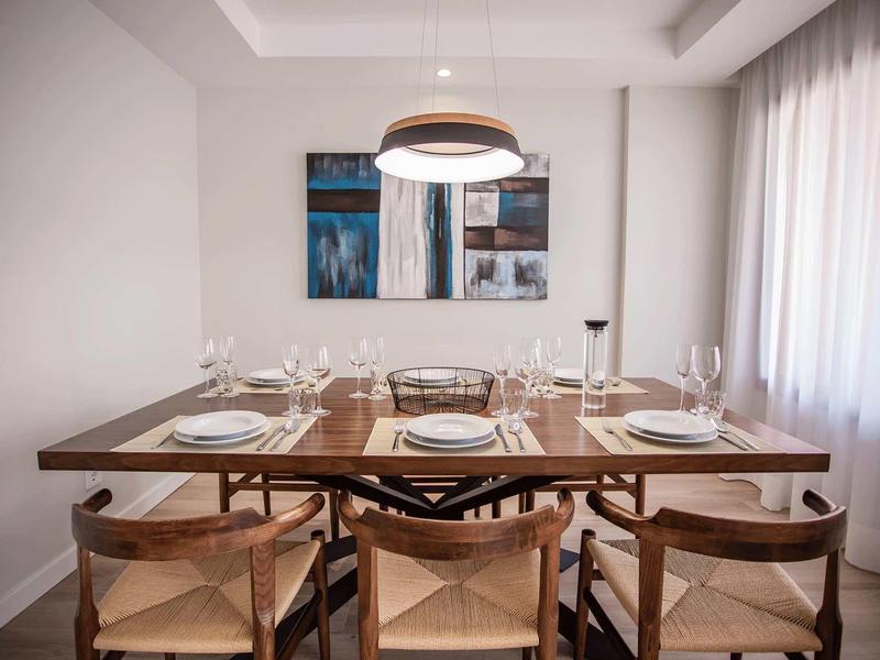 Mesa de comedor moderna con seis sillas, puesta con platos, copas y cubiertos en un espacio luminoso.