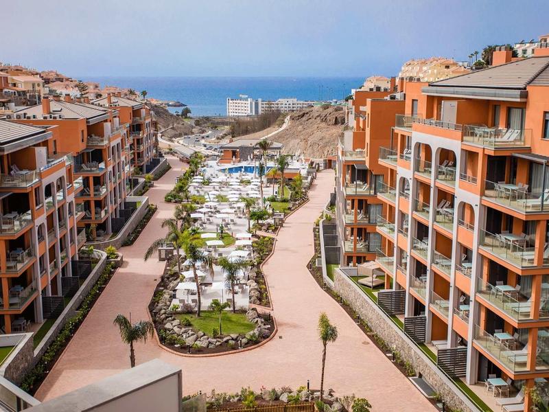 Vista de complejo hotelero animado con piscinas, caminos y edificios naranjas frente al mar.