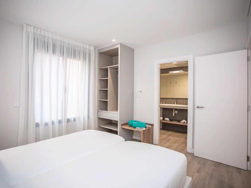 Dormitorio luminoso con cama blanca, ventana con cortinas y vista a baño con puerta abierta
