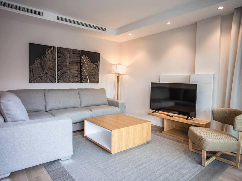 Sala moderna con sofá gris, mesa de madera, televisor y luces de acento en una habitación de hotel.