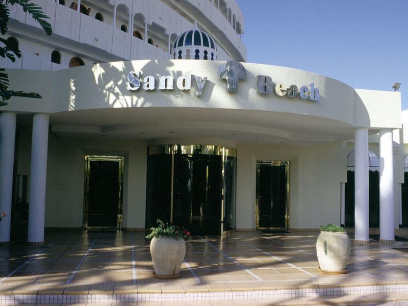 Entrée d'un hôtel blanc avec des colonnes rondes et des pots de fleurs devant.