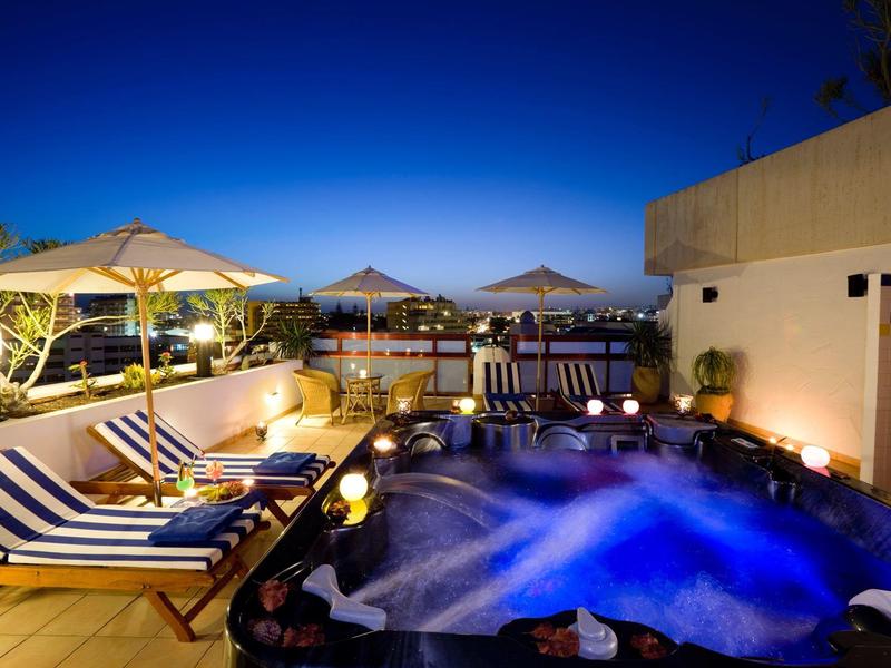Piscine d'hôtel éclairée en soirée avec chaises longues rayées et eau bleutée sous un ciel clair.