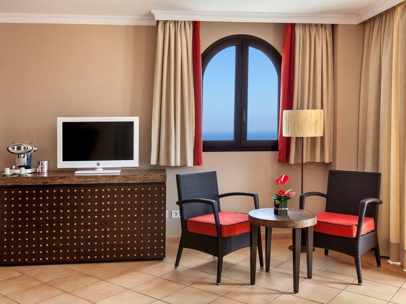 Coin salon d'hôtel avec deux fauteuils rouges, table et vue sur l'océan par une fenêtre arrondie.