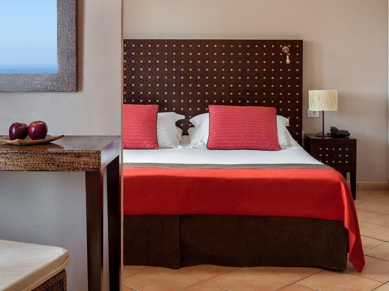 Chambre d'hôtel moderne avec lit double, couverture rouge et vue sur la mer depuis la fenêtre.