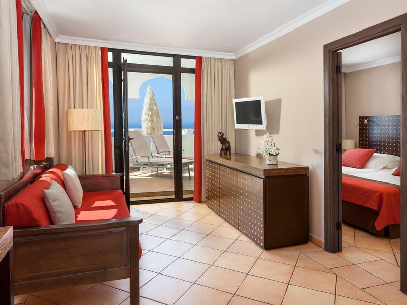 Appartement d'hôtel lumineux avec balcon, canapé rouge, télévision et chambre à lit rouge.