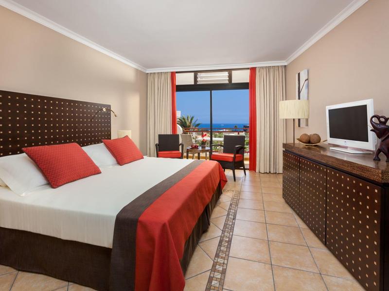 Chambre d'hôtel moderne avec lit double, couverture rouge, télévision et vue sur la mer depuis le balcon.