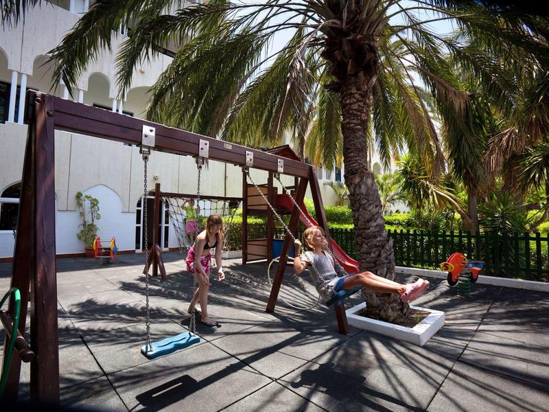 Aire de jeux avec balançoires sous des palmiers à côté d'un bâtiment blanc par temps ensoleillé