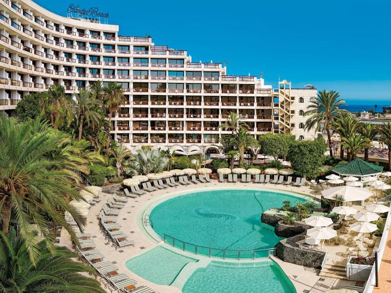 Hôtel avec piscine extérieure ronde, nombreuses chaises longues et palmiers sous un ciel clair.