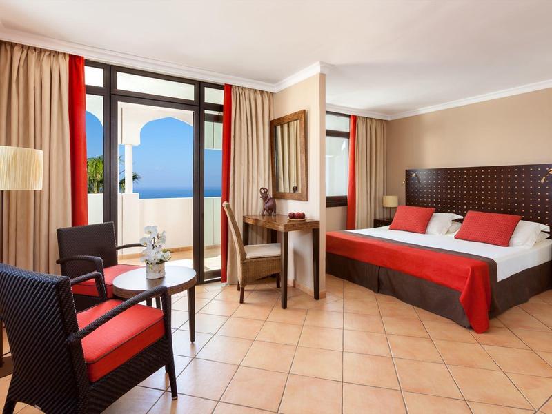 Chambre d'hôtel lumineuse avec lit double, décoration rouge et balcon avec vue sur la mer.