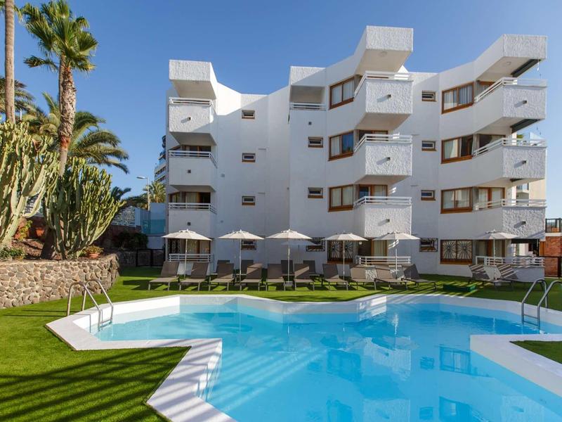 Moderní bílý hotel s bazénem a zeleným trávníkem pod jasnou oblohou.