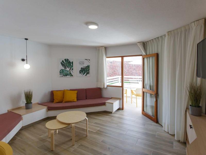 Obývací pokoj s rohovou pohovkou, kulatými stolky, rostlinami a televizí s přístupem na balkon.