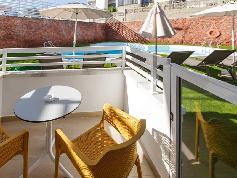 Malá terasa se stolem a žlutými židlemi vedle trávníku s lehátky a slunečníky.