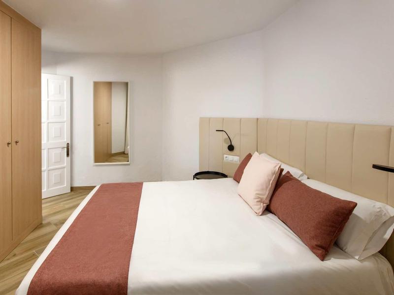 Moderní hotelový pokoj s manželskou postelí, dřevěnou skříní, velkým zrcadlem a minimalistickým nástěnným osvětlením.