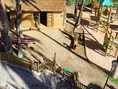 Children's playground with climbing frame, sandy ground, and wooden house in a hotel garden.