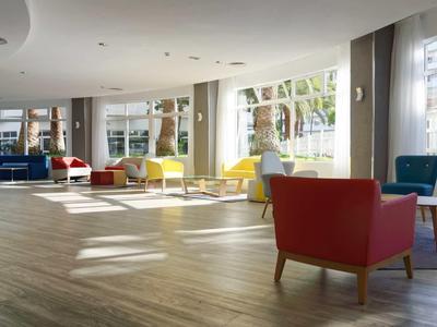 Spacious hotel lounge area with bright armchairs and large windows along the wall.