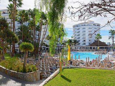 View of a hotel with pool, surrounded by palm trees and lounge chairs in sunshine.