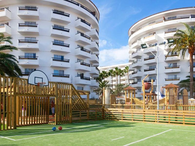 Modern basketball court in front of two round hotel buildings with palm trees under blue sky.