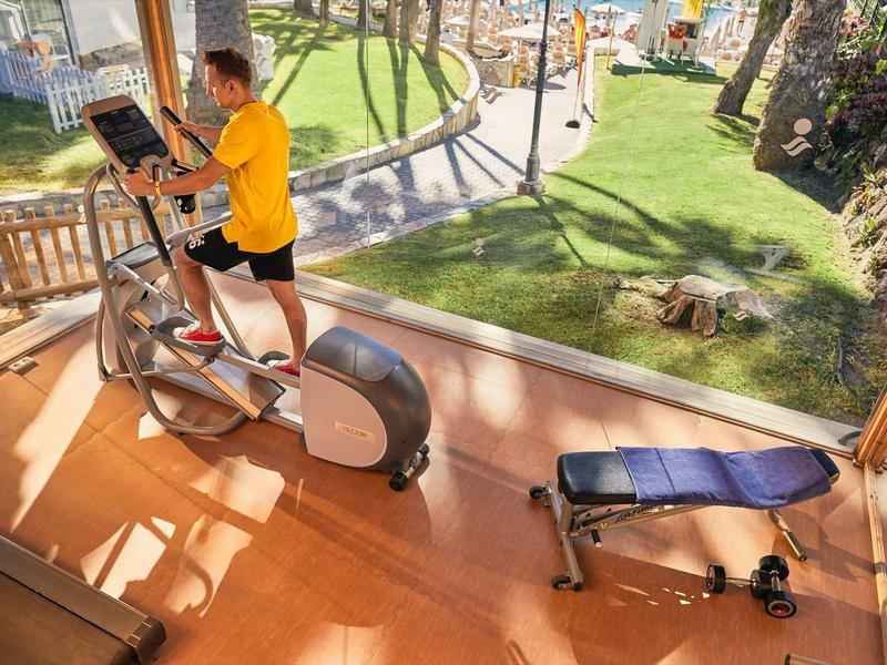 Man exercises on elliptical trainer in gym overlooking garden and pool.