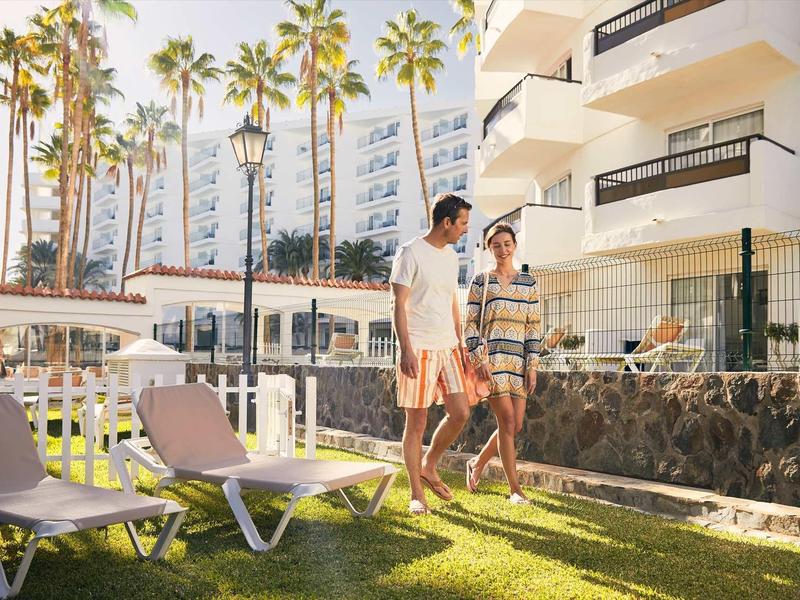 Couple walks leisurely on a lawn in front of an apartment hotel with palm trees.