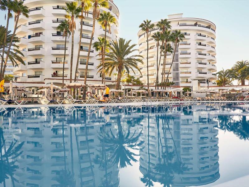 Modern white hotel buildings with palm trees and large clear swimming pool in the foreground.