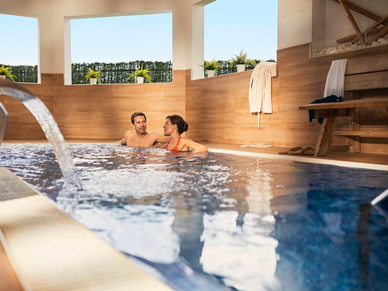 Couple relaxing in outdoor whirlpool with wooden paneling and water fountain under sunny weather.