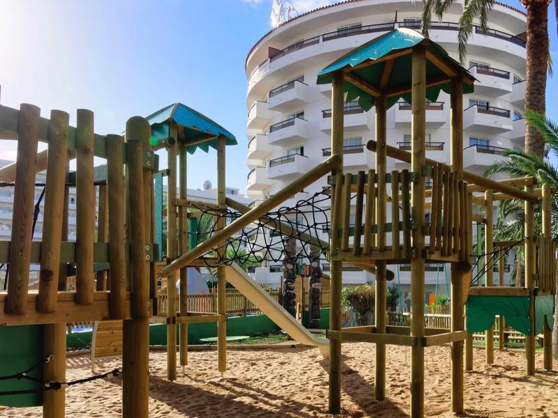 Playground with climbing nets and slides in front of a modern hotel building under blue sky.