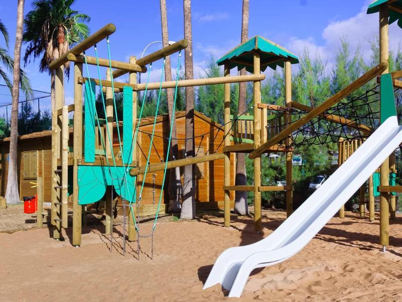 Playground with climbing frame, slide, and sandy ground under blue sky and palm trees.