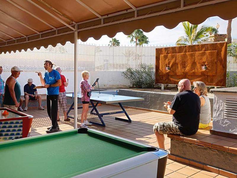 People playing table tennis and billiards in a sunny outdoor area with wooden canopy.