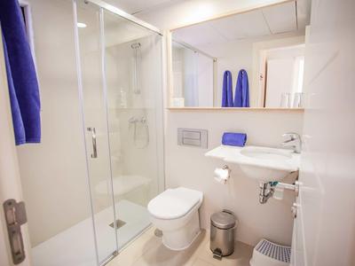 Kleines, helles Badezimmer mit Glas-Dusche, weißer Toilette, Waschbecken und blauen Handtüchern.