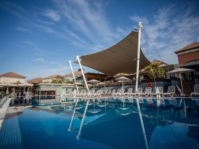 Großer blauer Pool mit Liegestühlen und Sonnensegeln, umgeben von Gebäuden unter blauem Himmel.