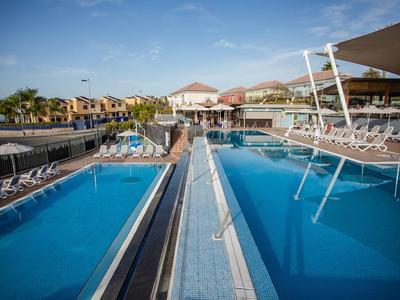 Zwei große Schwimmbecken mit blauen Liegestühlen und Palmen unter blauem Himmel in einem Resort.