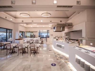 Weitläufiger Speisesaal mit weißen Tischen, Stühlen und großen Fenstern, heller Marmorboden.