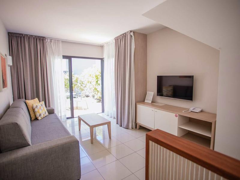 Helles Wohnzimmer mit grauem Sofa, kleinem Tisch, TV an der Wand und großen Glastüren zum Balkon.