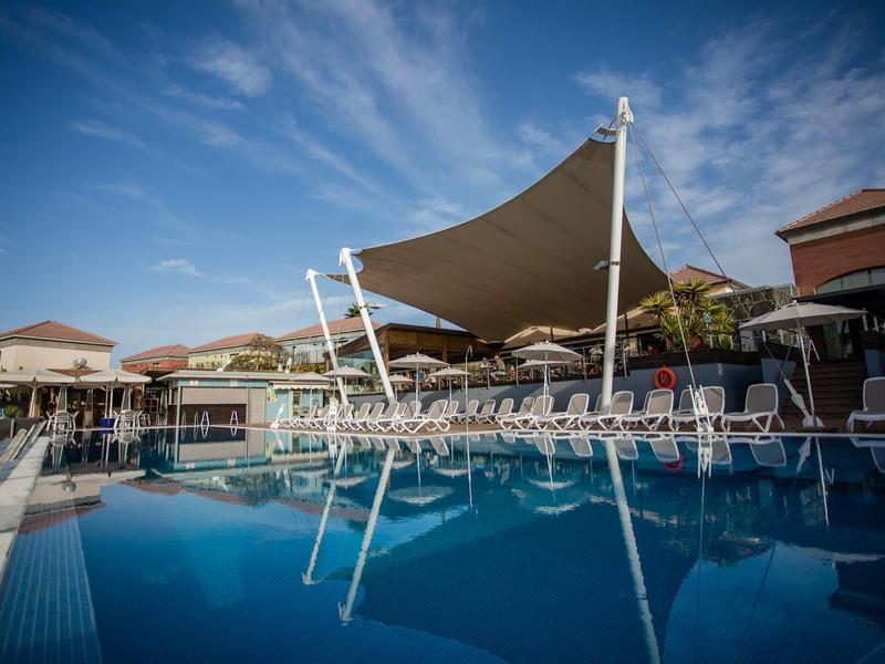 Großer blauer Pool mit Liegestühlen und Sonnensegeln, umgeben von Gebäuden unter blauem Himmel.