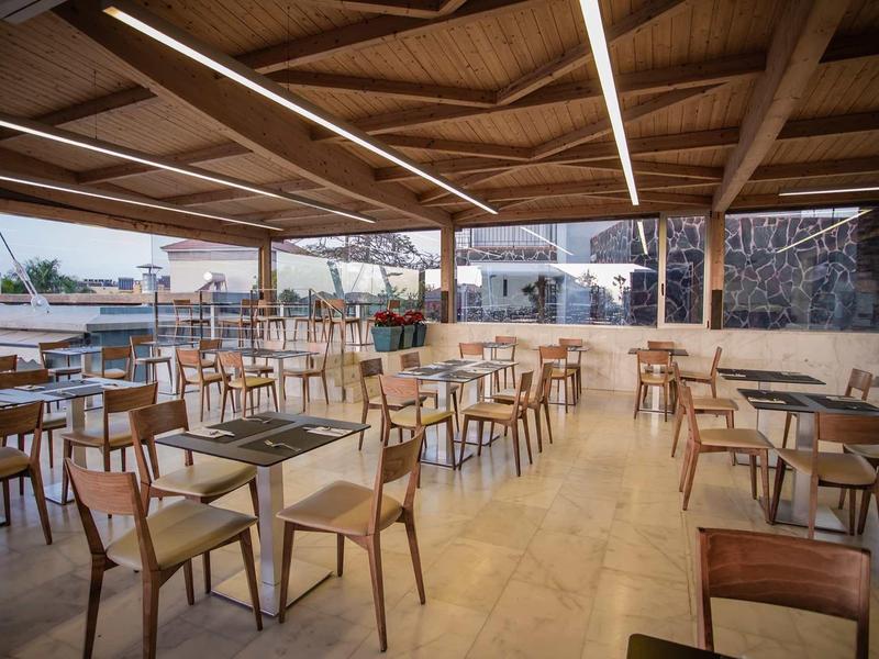 Offene Terrasse mit Holzdecke, vielen Tischen und Stühlen, heller Bodenbelag, Blick auf Wasser und Stadt.