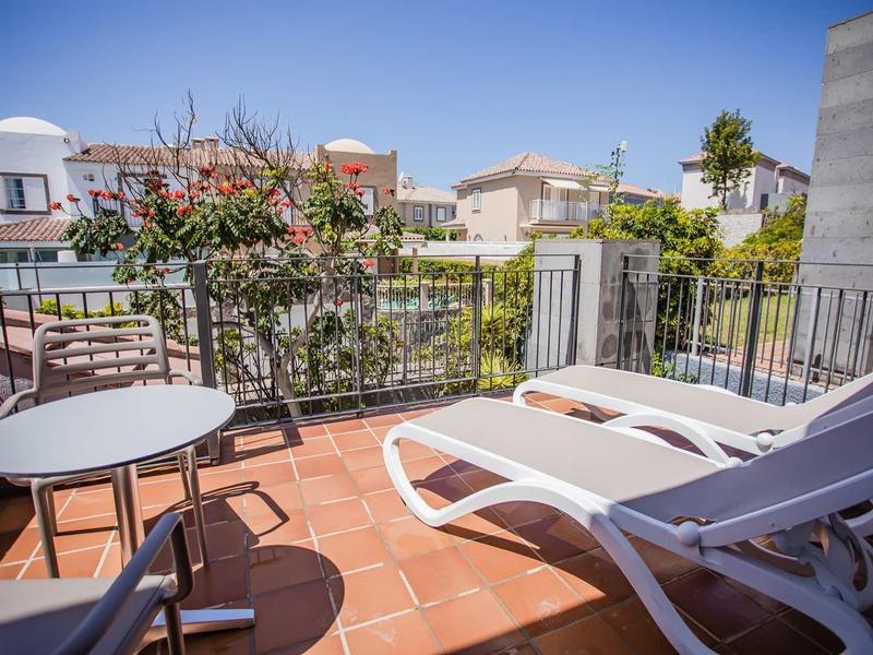 Terrasse mit roten Fliesen, zwei weißen Liegestühlen, kleinem runden Tisch und Stühlen, sonnig und klar.