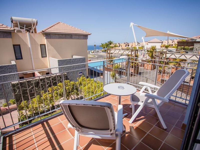 Balkon mit weißen Stühlen und Tisch, Blick auf Wohnhäuser, Pool und blauen Himmel.