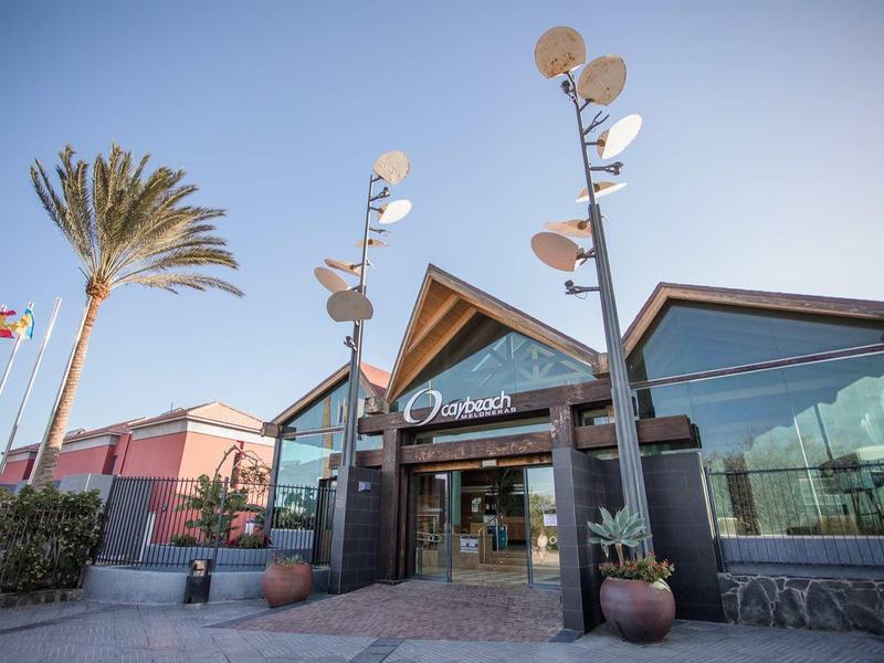 Eingang eines modernen Gebäudes mit Glasfront, Palmen und klar blauem Himmel im Hintergrund.