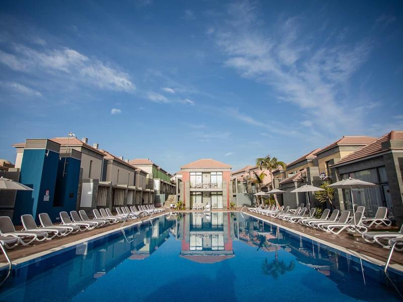 Großer, klarer Pool mit Liegestühlen, umgeben von modernen, zweistöckigen Wohngebäuden unter blauem Himmel.