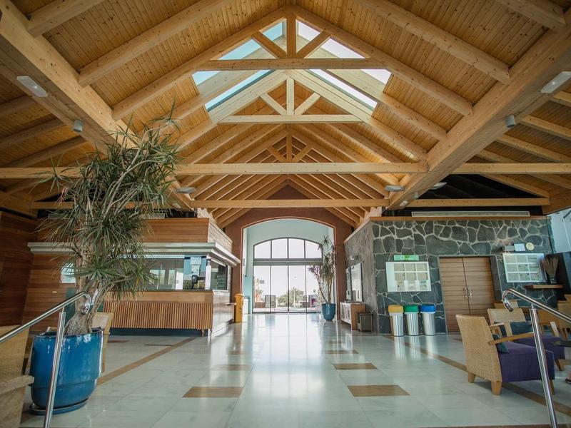 Großes Holzgebäude mit transparentem Dach, Sitzbereich und Pflanzen im Eingangsbereich.