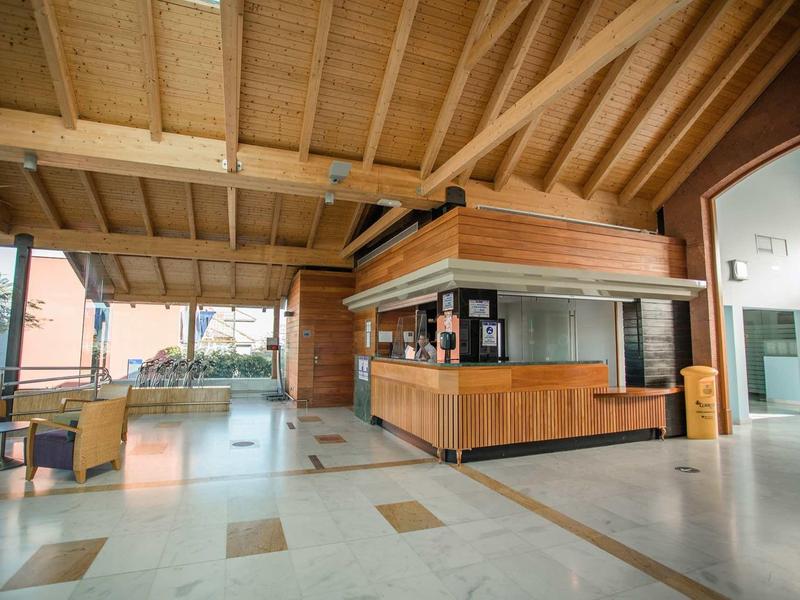 Großer Raum mit Holzbalkendecke, Empfangstheke aus Holz und Glas, Sitzbereich und großem Fenster.